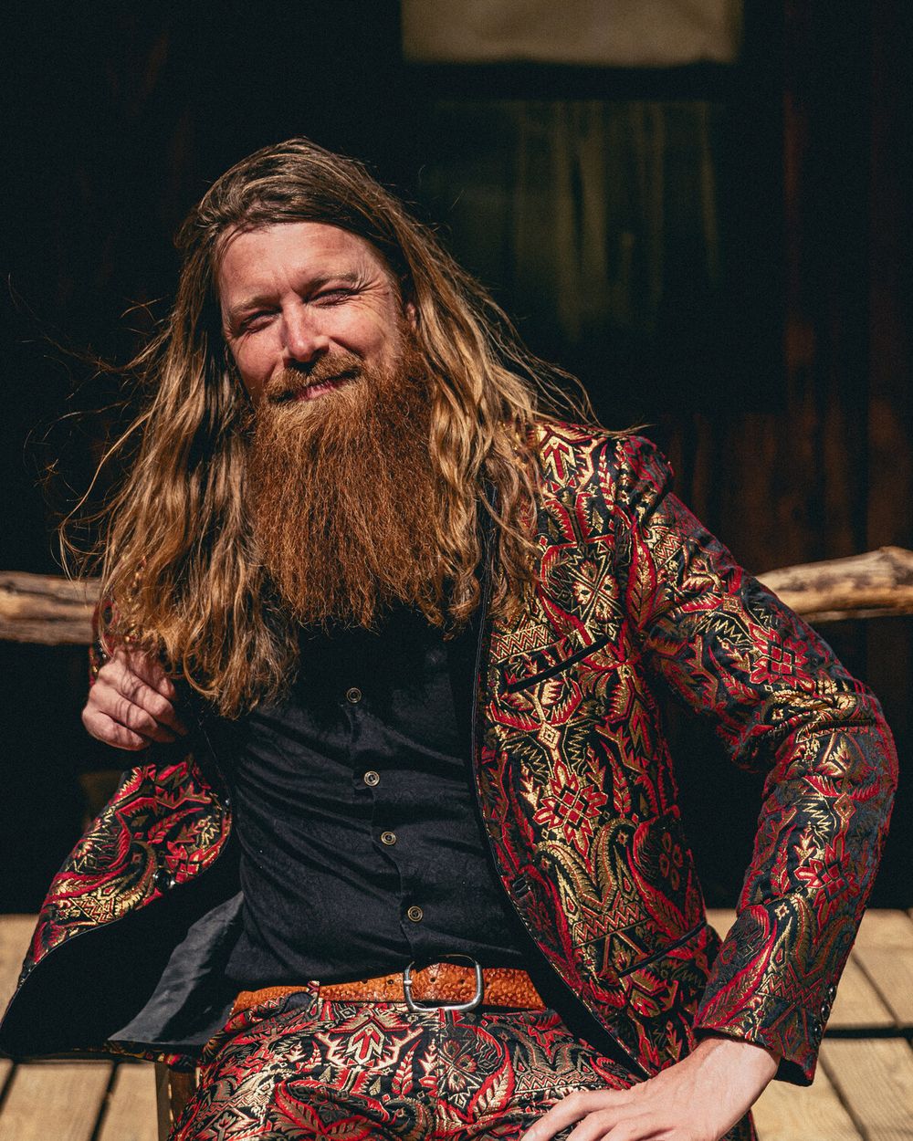 Shane Mauss, bearded and long-haired in a red-and-gold brocade suit, squinting into the sun
